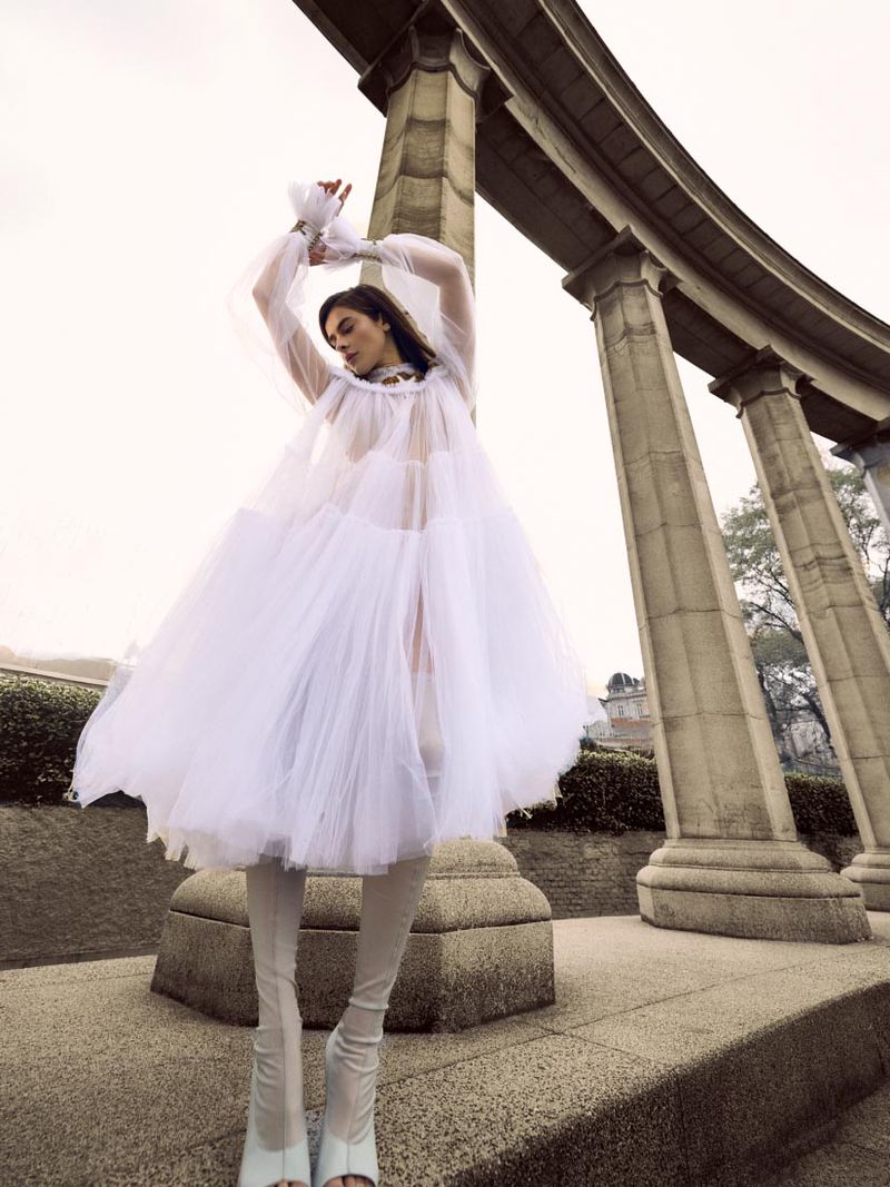 Woman in tulle dress posing in front of concrete columns, architectural details, and overcast sky, capturing a fashion-forward editorial.
