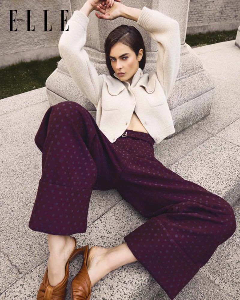 Fashion editorial shot of a woman in a cream-colored sweater and burgundy pants, seated on a stone surface with her arms raised.