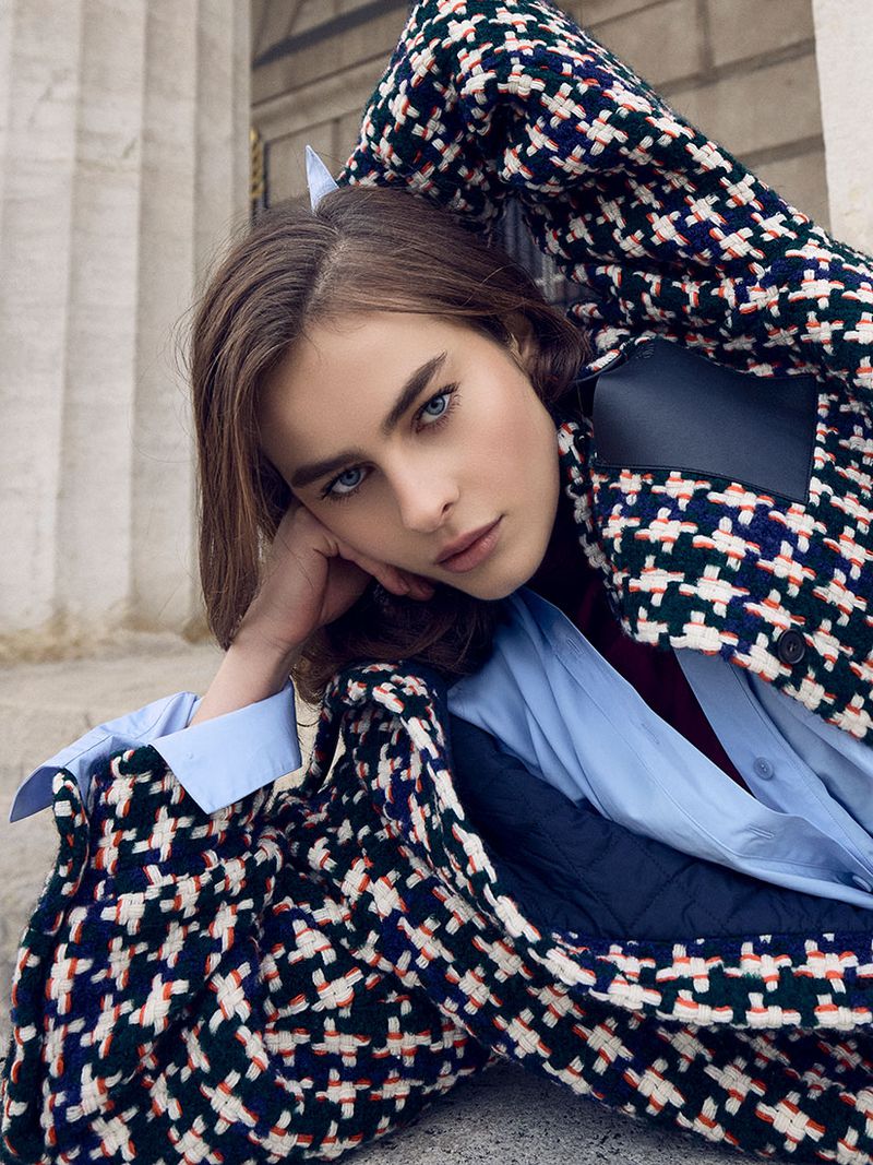 A young woman with blue eyes poses with a patterned jacket in an outdoor setting featuring a stone staircase and Roman-style columns.