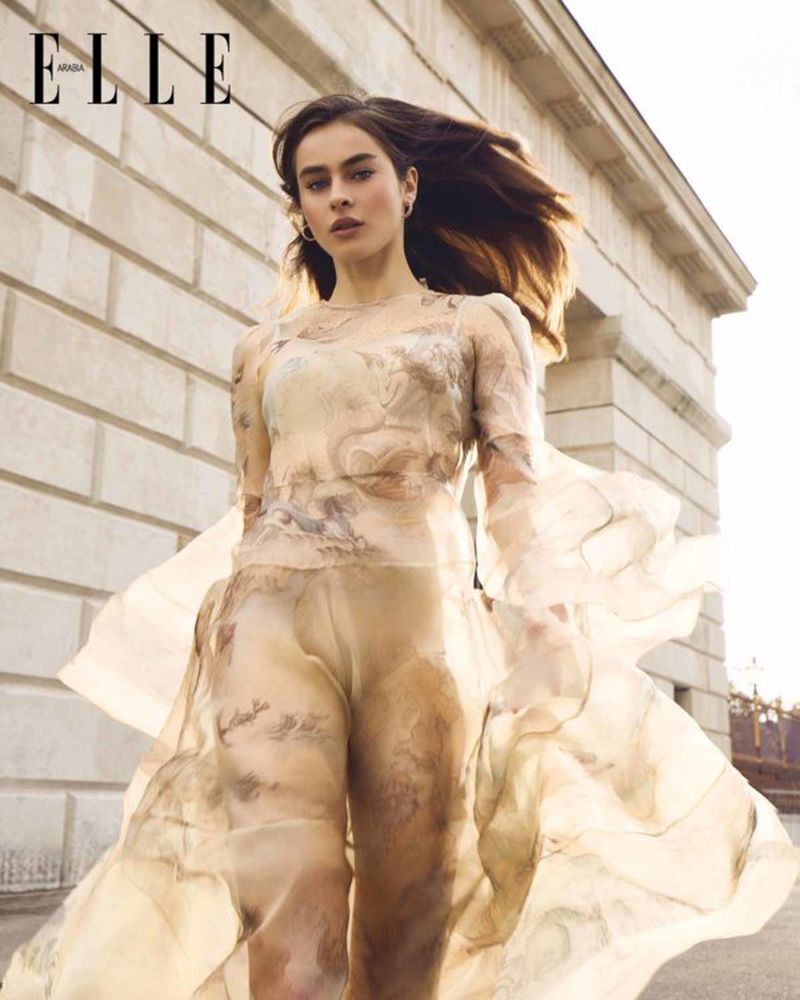 Fashion editorial featuring a young woman in a sheer, patterned dress standing outdoors with a stone building in the background. 