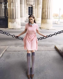 A woman in a pink dress and grey knee-high socks poses between neoclassical columns holding a black chain