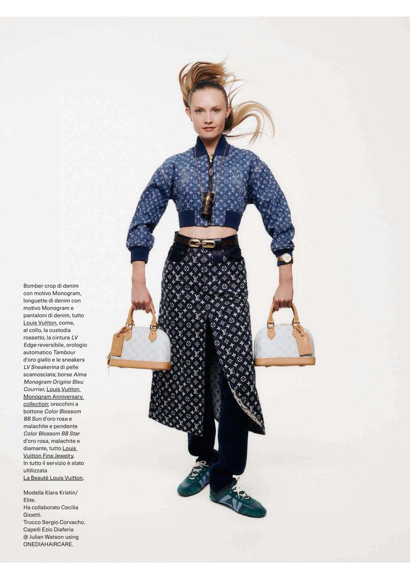 A blonde female fashion model poses in a patterned denim jacket and skirt, holding matching purses against a solid neutral backdrop.
