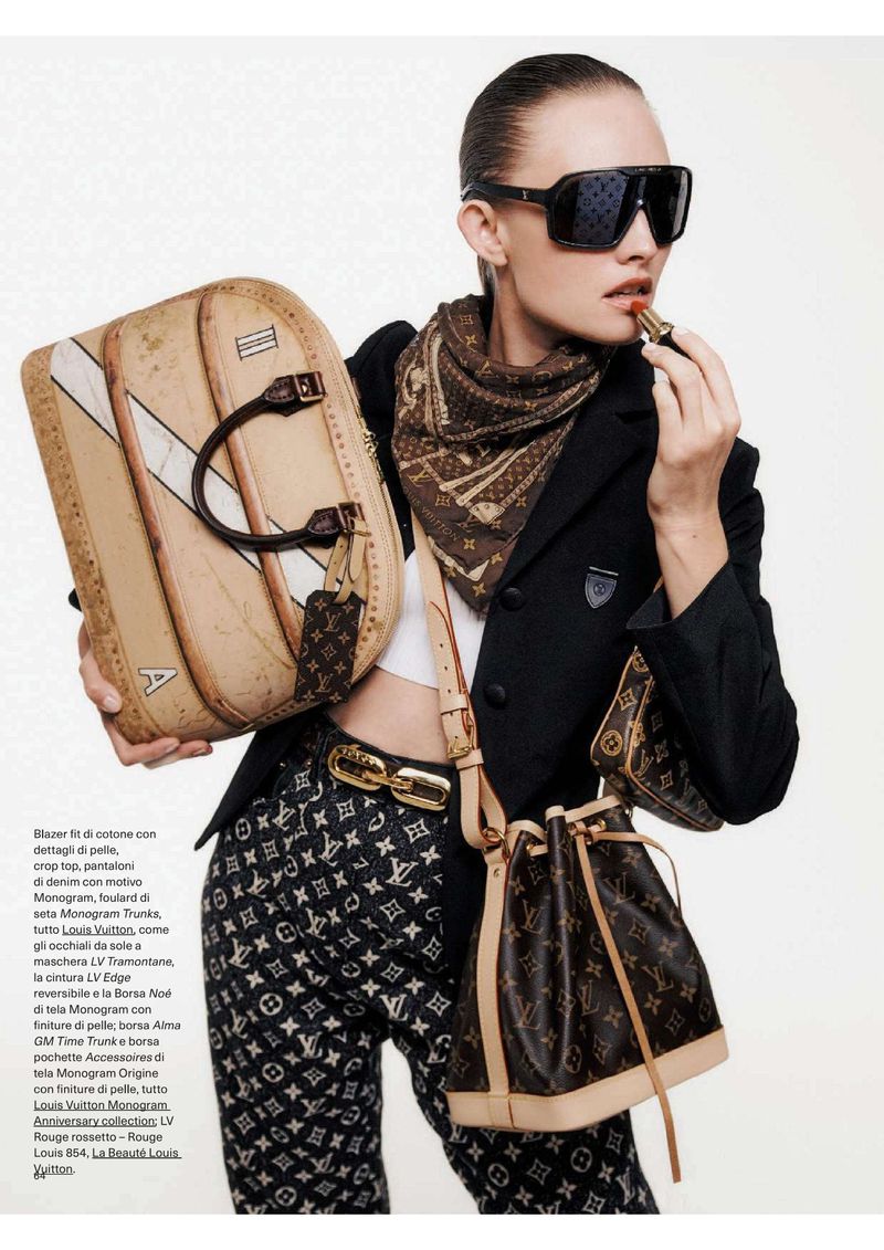 Fashionable woman applying lipstick while holding tan suitcase and wearing black blazer, patterned pants, and Louis Vuitton accessories.