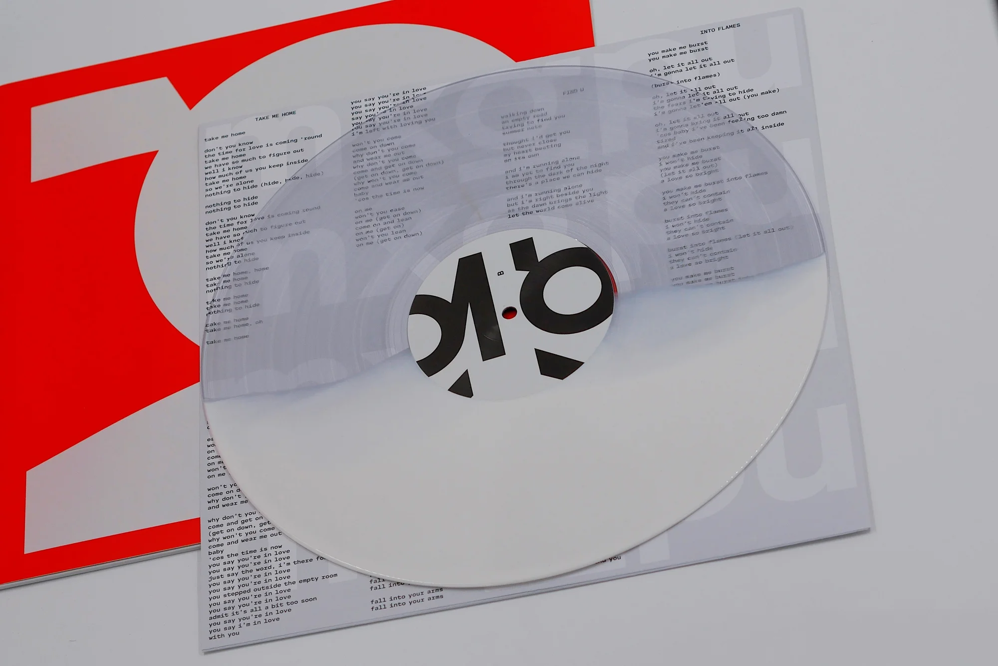 Image displays a clear vinyl record on a cover with lyrics, next to an album sleeve featuring bold red and white text.