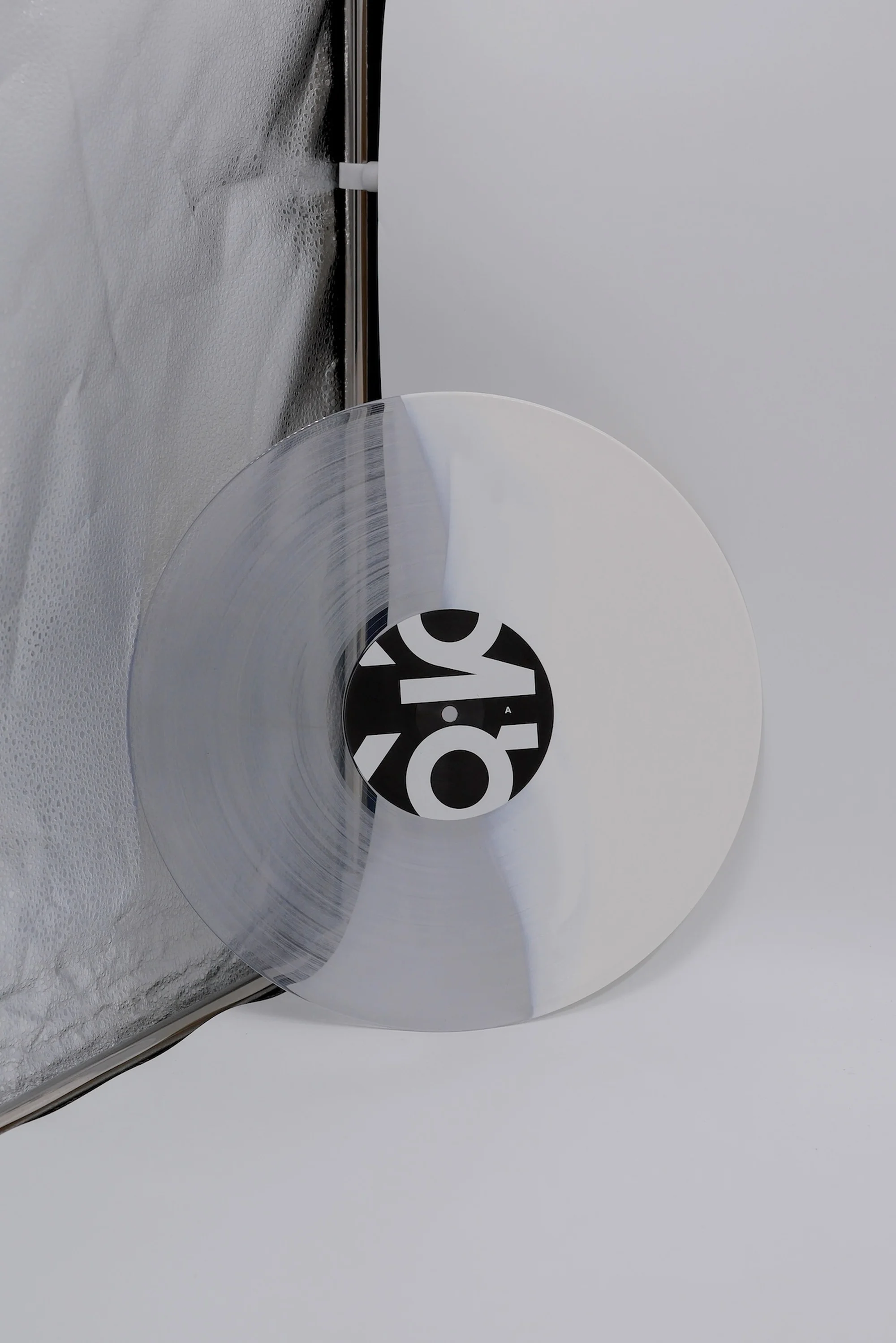 Clear and white vinyl record with a black and white label, presented against a crinkled silver and white backdrop in a studio setting.