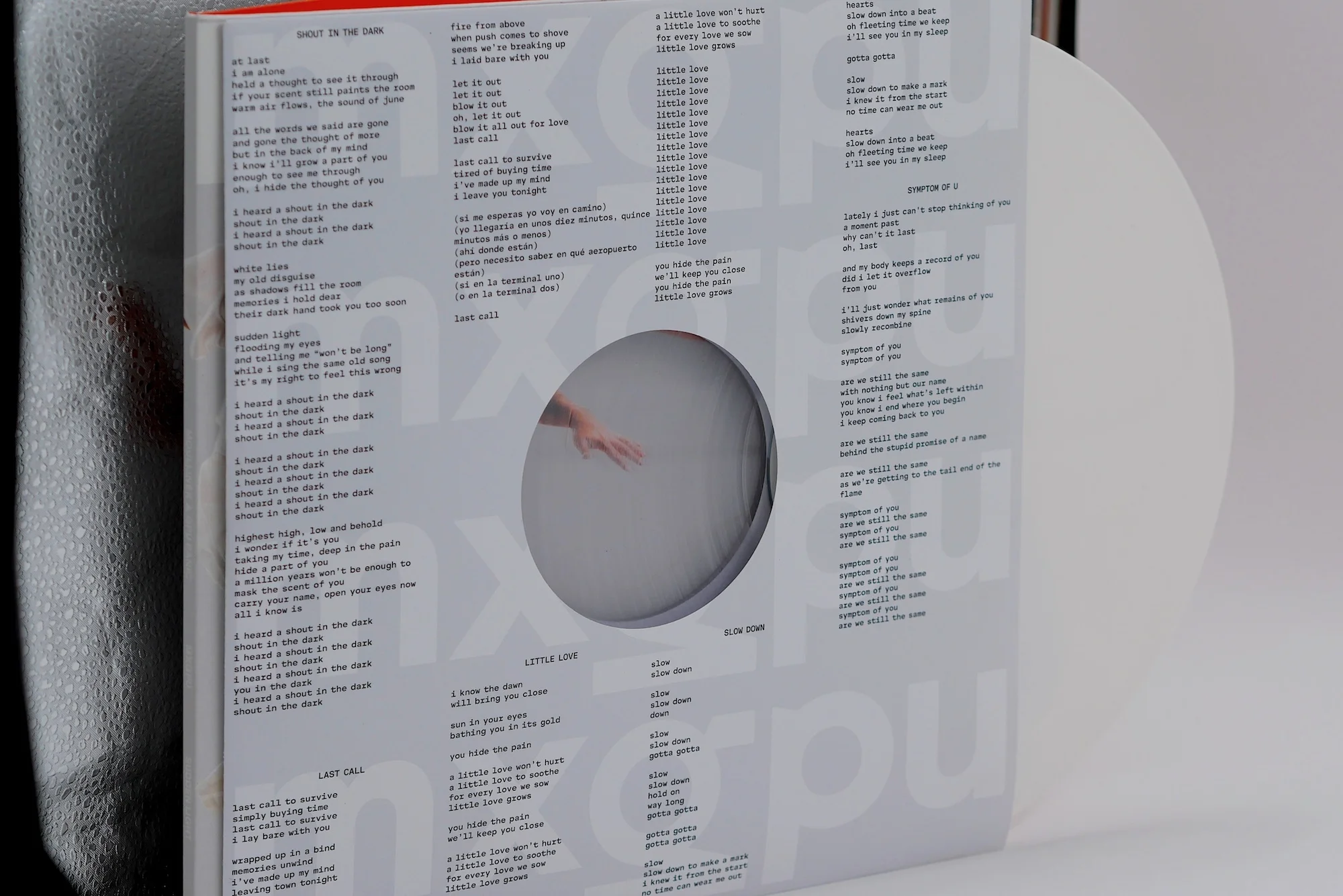 Record sleeve with printed lyrics featuring a cutout revealing a glimpse of a human hand, against a white backdrop. 
