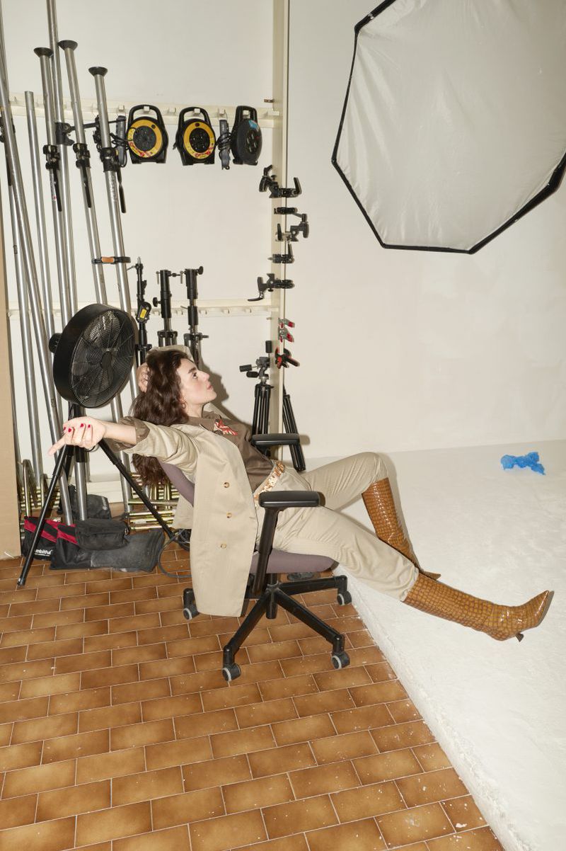 Stylish woman in a tan suit and knee-high boots poses in a studio on a rolling chair, surrounded by photography equipment.
