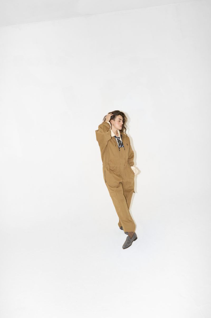 A woman with curly brown hair poses in a tan jumpsuit and grey shoes against a white studio backdrop. She is running a hand through her hair.