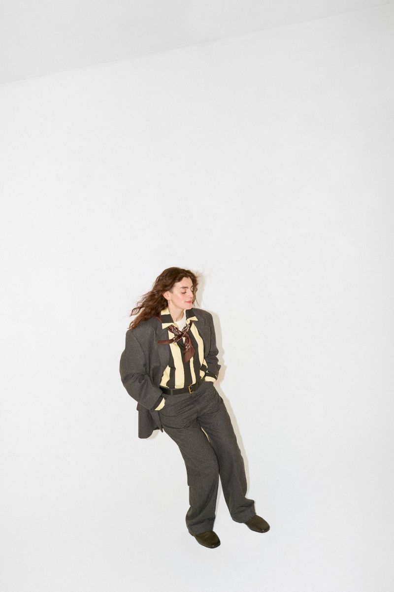 A young woman with curly brown hair poses in a gray suit and striped shirt, shot in a bright photo studio with ample white space.