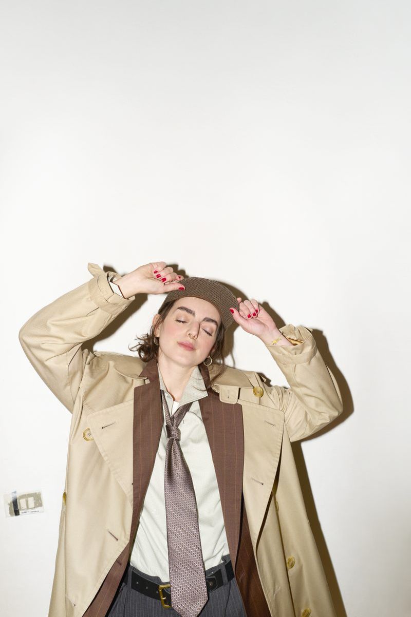 A woman in a tan trench coat and beret poses in front of a plain white wall with her eyes closed and her hands near her head.