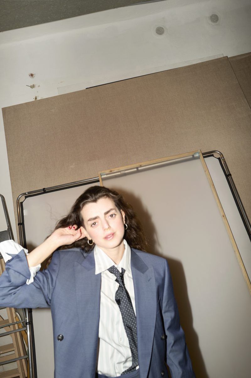 Woman in blue suit and tie strikes a confident pose indoors, exuding modern professional style with neutral background and soft lighting.