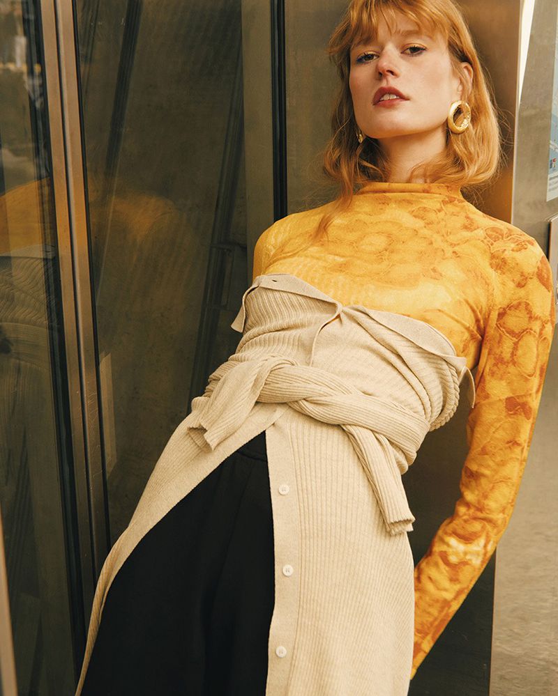 Portrait of a red-haired woman in a patterned yellow top and beige ribbed cardigan, posing against a textured wall with a neutral expression.