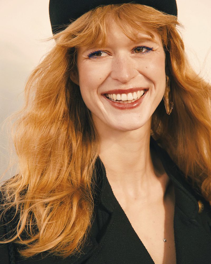 Close-up portrait of a smiling red-haired woman wearing a black beret and dark jacket against a plain background.