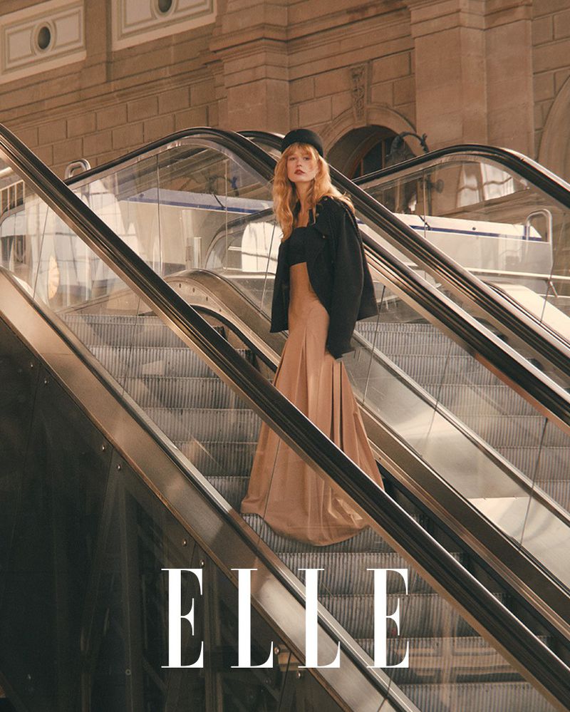Woman in black jacket and tan skirt poses on escalator in urban setting with stone architecture and ELLE magazine logo overlay.