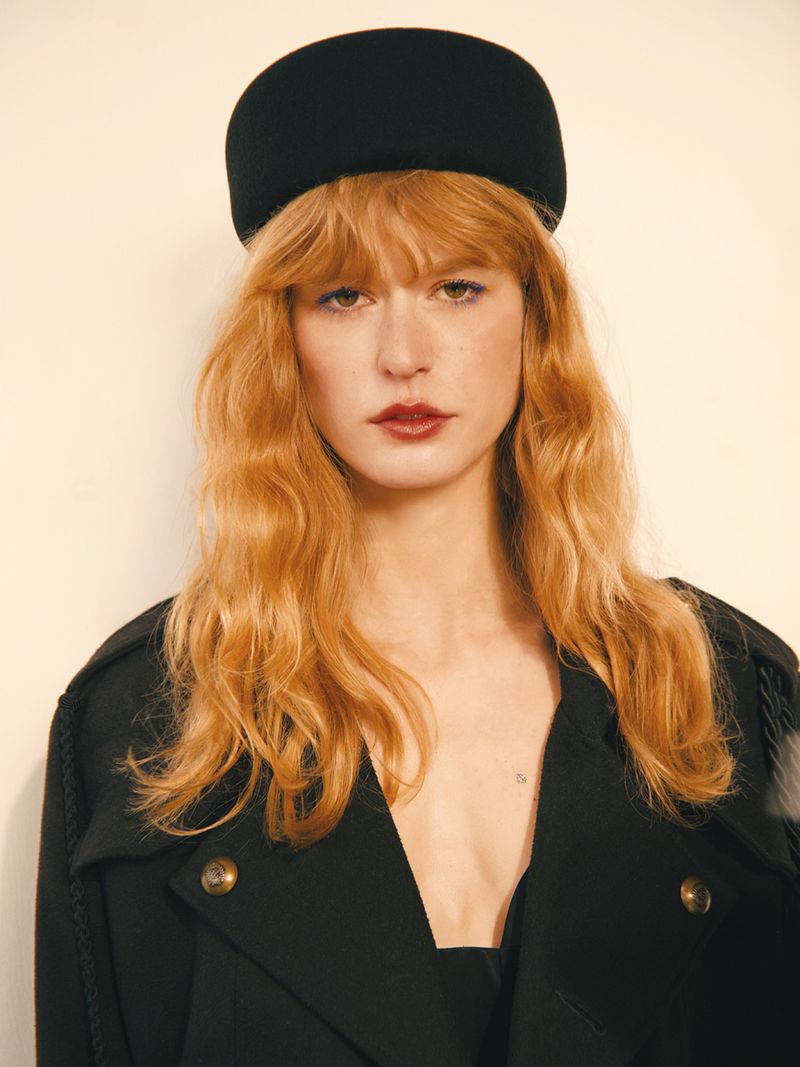 Fashion editorial shot of a woman with dark hair wearing a black outfit and posing in front of a textured gold background.