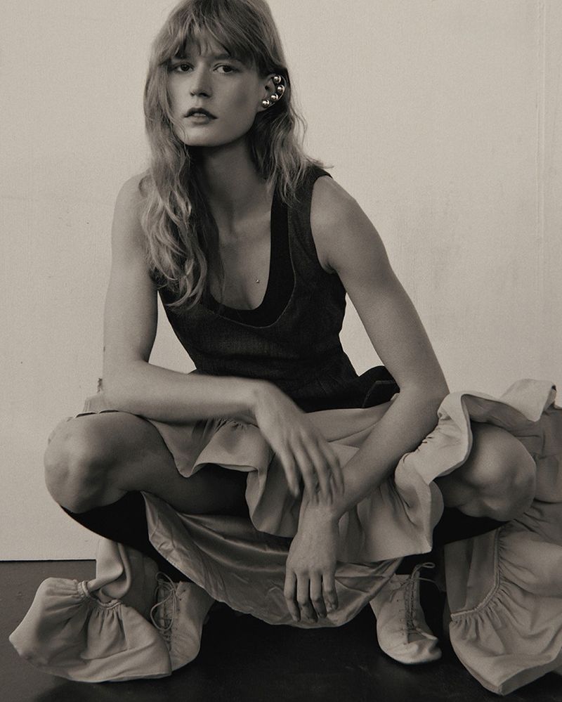 Monochrome studio shot of a blonde woman squatting, wearing a dark top and ruffled skirt, posing with a serious expression.