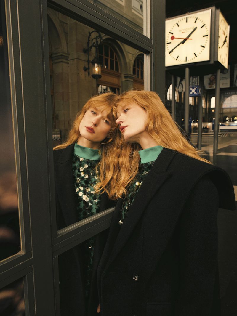 Adult woman with long red hair, wearing a black coat and sequined shirt, poses next to window with clock in blurred background at night.