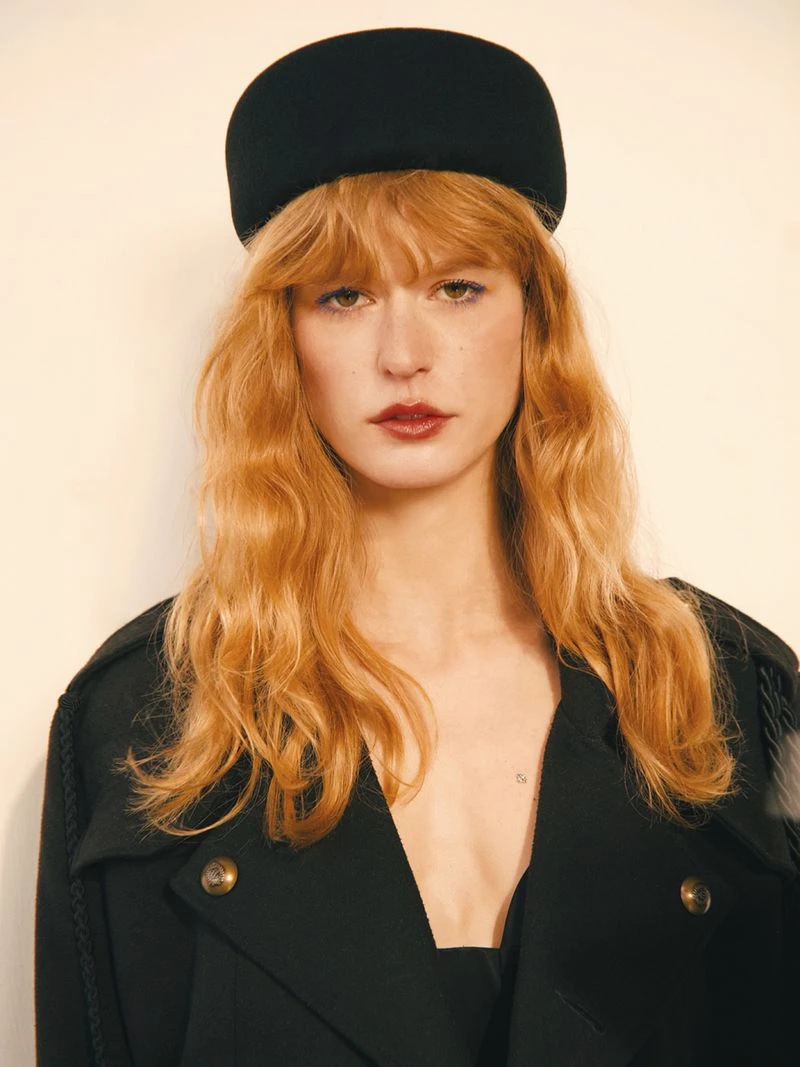 Model with long red hair wearing a dark hat and black coat against a neutral background for Elle Serbia editorial