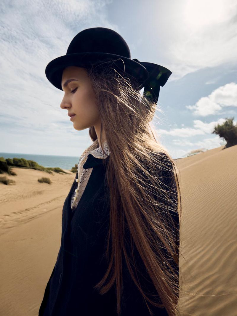 Woman in a black bowler hat poses in profile on a sand dune with the ocean and a cloudy sky in the background on a sunny day.