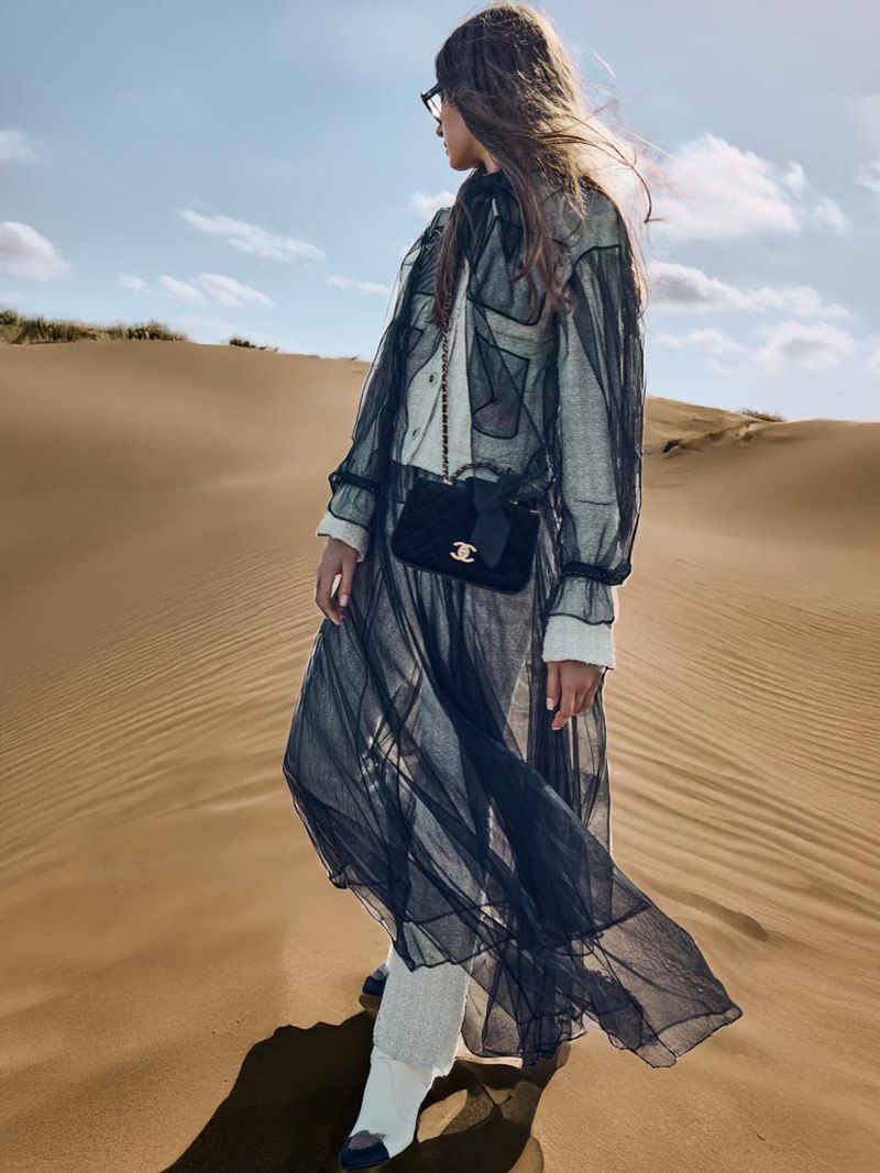 Fashion shot of a woman in a sheer black dress and white pants, standing on a sand dune under a bright blue sky with scattered clouds.