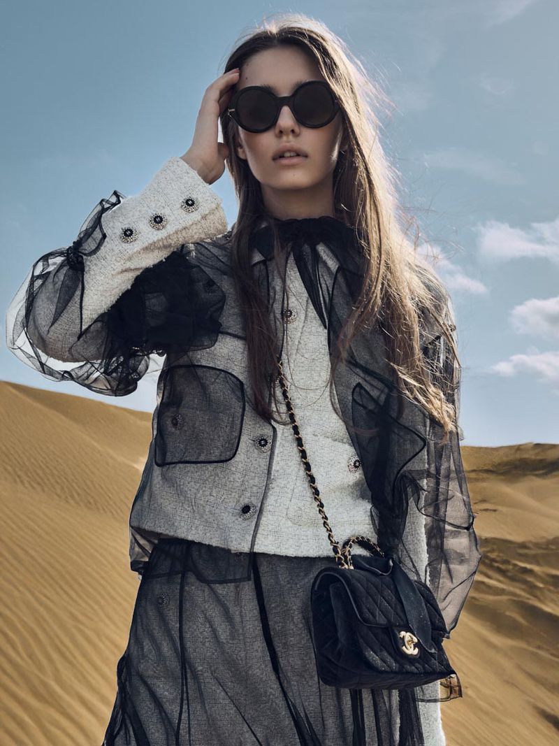 Fashionable woman in sunglasses and designer coat poses in a desert landscape with blue sky, showcasing a luxury handbag.