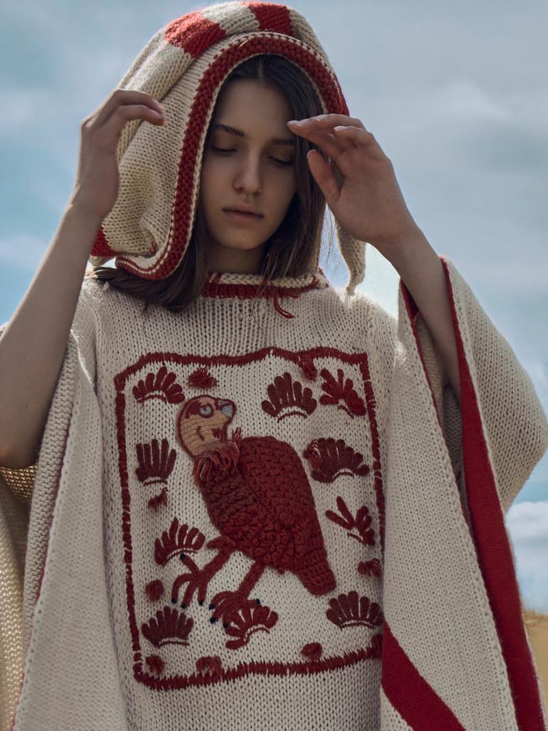 Young woman in a knit poncho with a hood, posing outdoors in a field. The poncho features a design of a stylized bird.