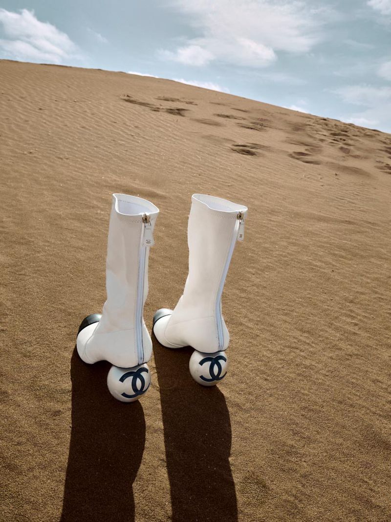 Fashionable women's white high-heel boots with black accents, featuring a Chanel logo on the heel, set against a desert landscape.