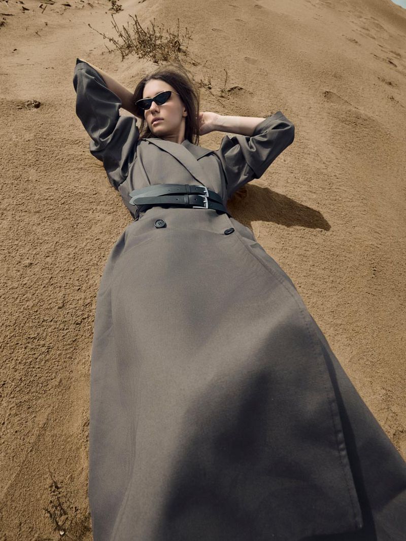A woman in a coat and sunglasses lies on a sand dune, hands behind her head. Desert landscape with neutral colors and a fashion editorial style.