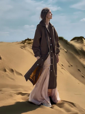 A woman in a long dress and coat stands in a desert landscape under a blue sky with clouds. She is looking off into the distance.