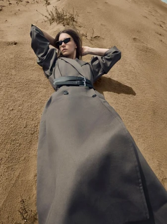 Model lying on sandy dunes wearing a long grey belted coat and sunglasses