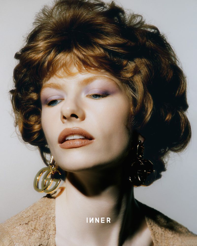 Close-up portrait of a stylish young woman with auburn hair, light lilac eyeshadow, nude lipstick, and gold earrings against a neutral background.