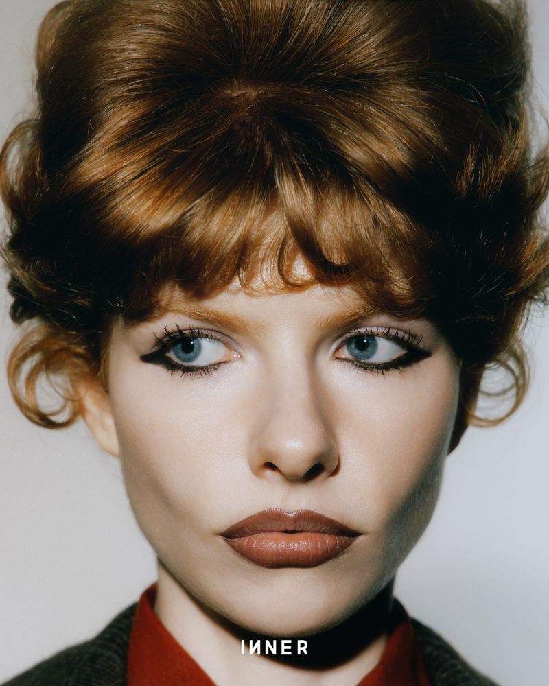 Close-up portrait of a woman with red hair and blue eyes wearing eyeliner and red lipstick looking off to the side in a studio setting.