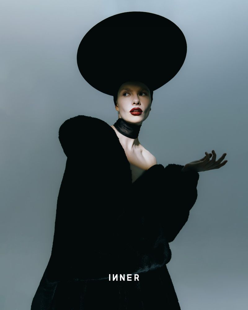 A woman with pale skin and dark red lipstick poses in a black sculptural coat and large hat against a gray studio backdrop.