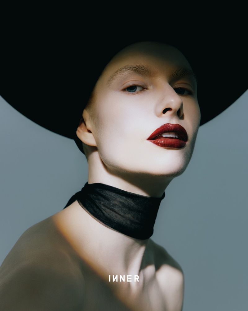 Glamour headshot of an adult female model in a wide-brimmed hat and black neck scarf with dark red lipstick against a grey backdrop.