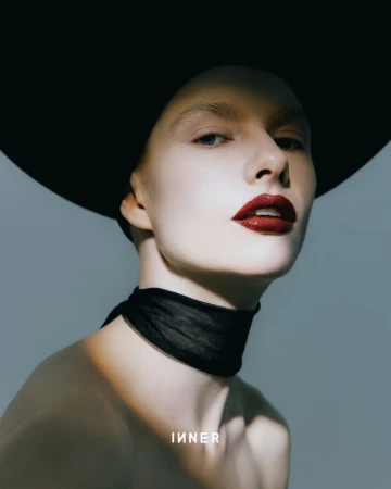 Glamour headshot of an adult female model in a wide-brimmed hat and black neck scarf with dark red lipstick against a grey backdrop.