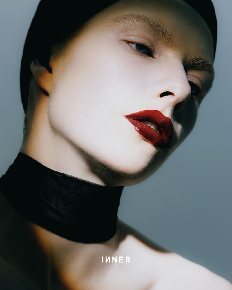 Close-up portrait of a light-skinned female model with red lipstick and black head covering, wearing a black choker against a gray backdrop.