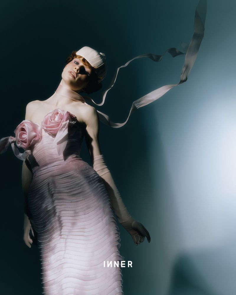 A model in a pink floral gown and white hat poses indoors against a blue background with ribbons, creating an elegant fashion image.