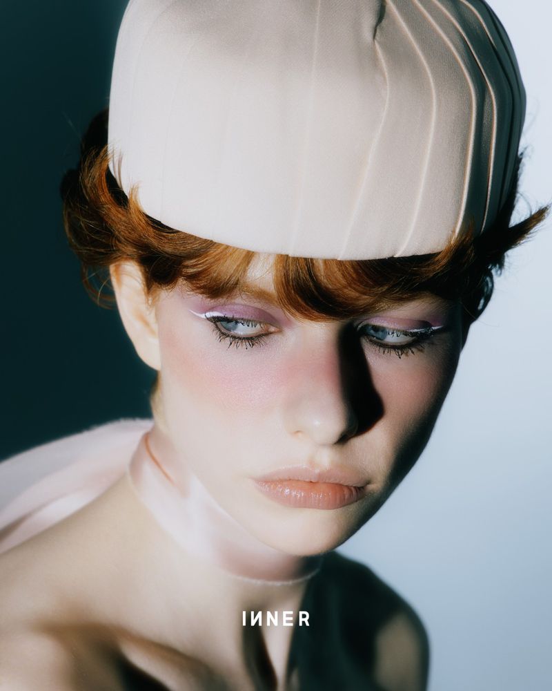 Close-up portrait of a young adult woman with red hair wearing a hat and light makeup, featuring soft, diffused lighting and a serene expression.