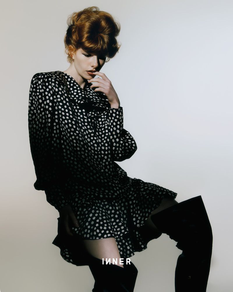 A red-haired woman poses in a black-and-white polka-dot dress and thigh-high boots against a plain backdrop for a fashion editorial.