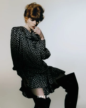 A red-haired woman poses in a black-and-white polka-dot dress and thigh-high boots against a plain backdrop for a fashion editorial.