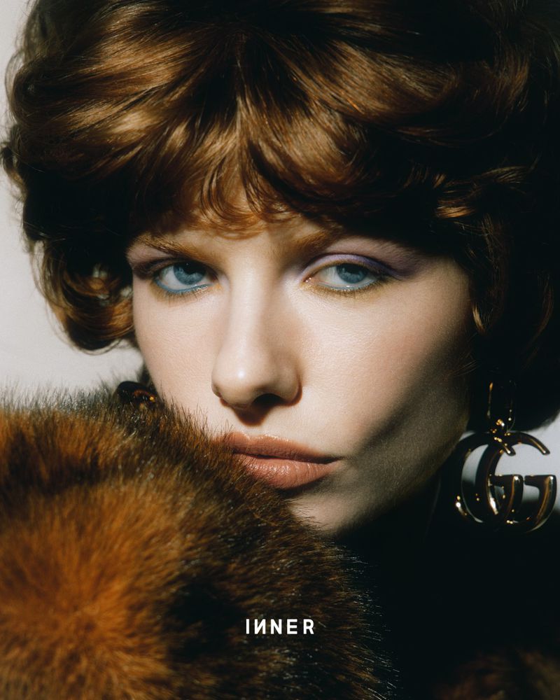 Close-up fashion portrait of a young adult woman with red hair, blue eyes, and Gucci earrings posing with a fur stole in a studio.