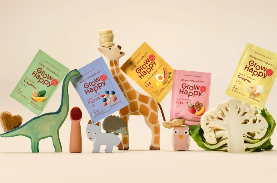 Assortment of Grow Happy allergen-immunobutter packets displayed among wooden children's toys and a halved cauliflower on a light surface.
