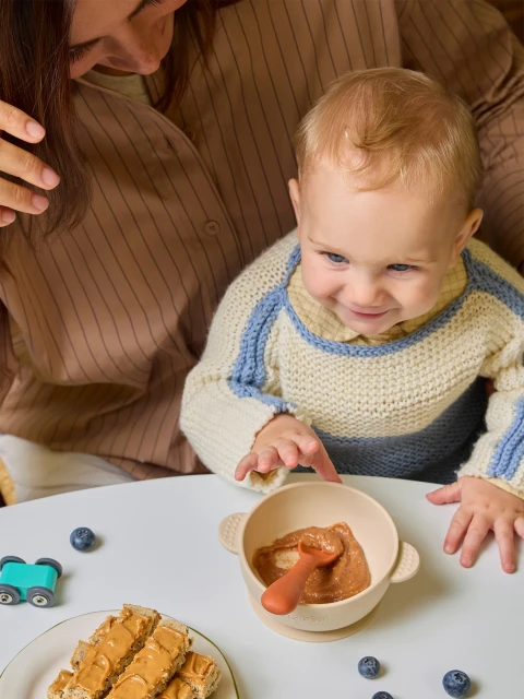 Blond baby smiles while reaching for food in a bowl on a table, with a woman's hand and various food items visible nearby.
