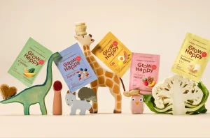 Colorful GrowHappy food pouches arranged among wooden animal figurines and fresh produce on a neutral background