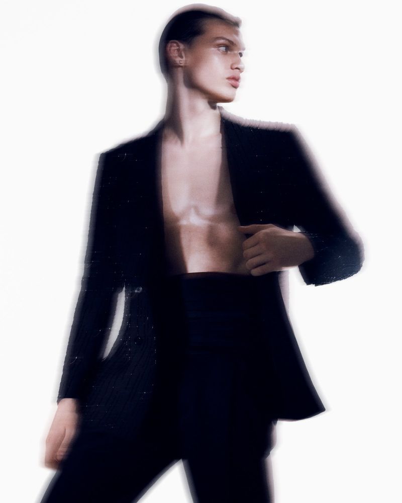 Portrait of a shirtless man in a black suit looking left, against a diffused white backdrop with bright light, minimal studio fashion look.