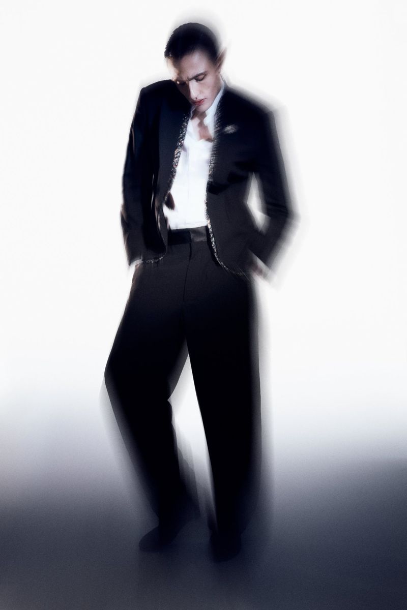 An adult male model posing in a black suit jacket, white shirt, and black pants with a white background creating an editorial fashion shot.