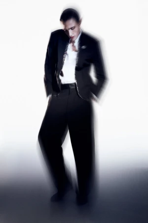 An adult male model posing in a black suit jacket, white shirt, and black pants with a white background creating an editorial fashion shot.
