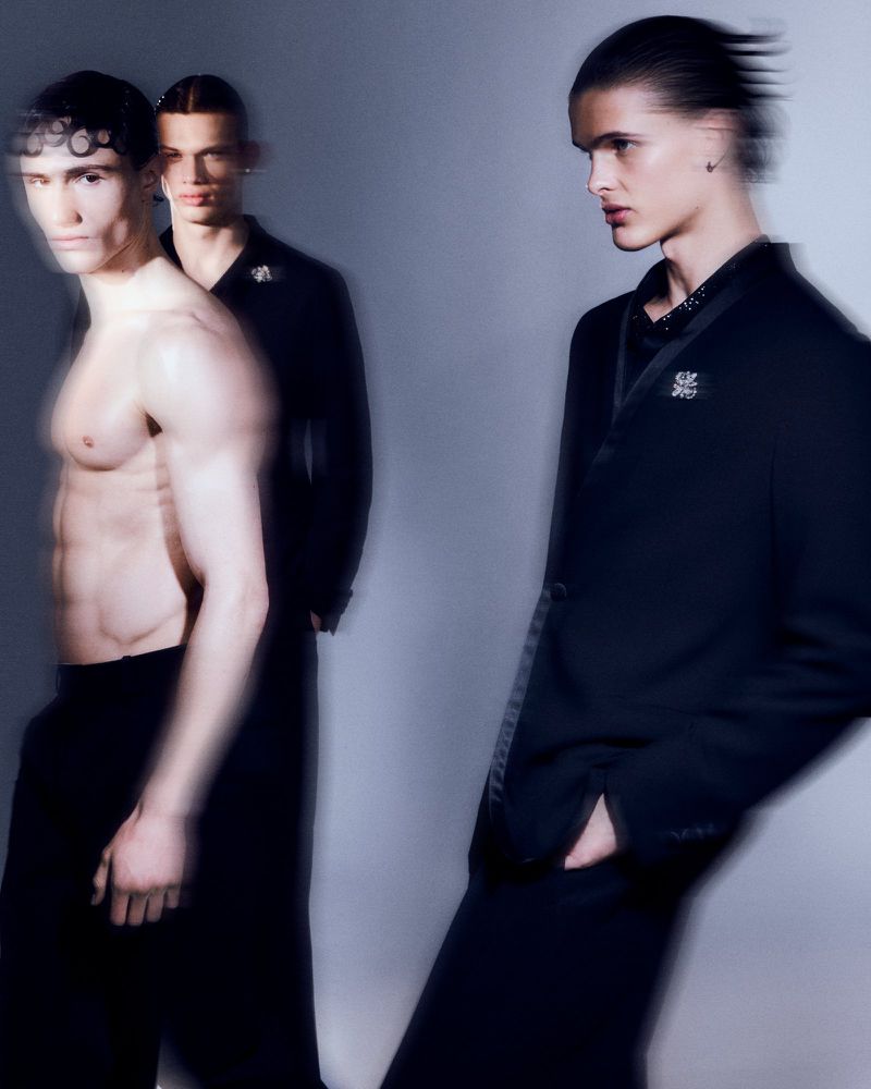 Three male fashion models in black suits and pants are featured in a blurred studio shot against a light gray background. 