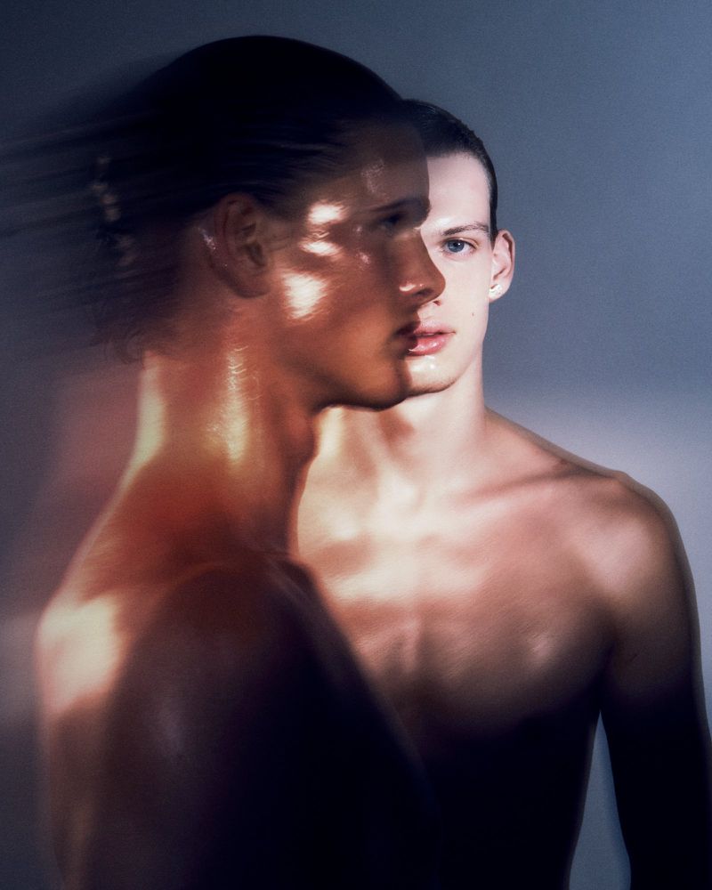 Striking studio portrait of two shirtless adult men with clean-shaven heads. One man is blurred in motion and contrasted against a grey backdrop.