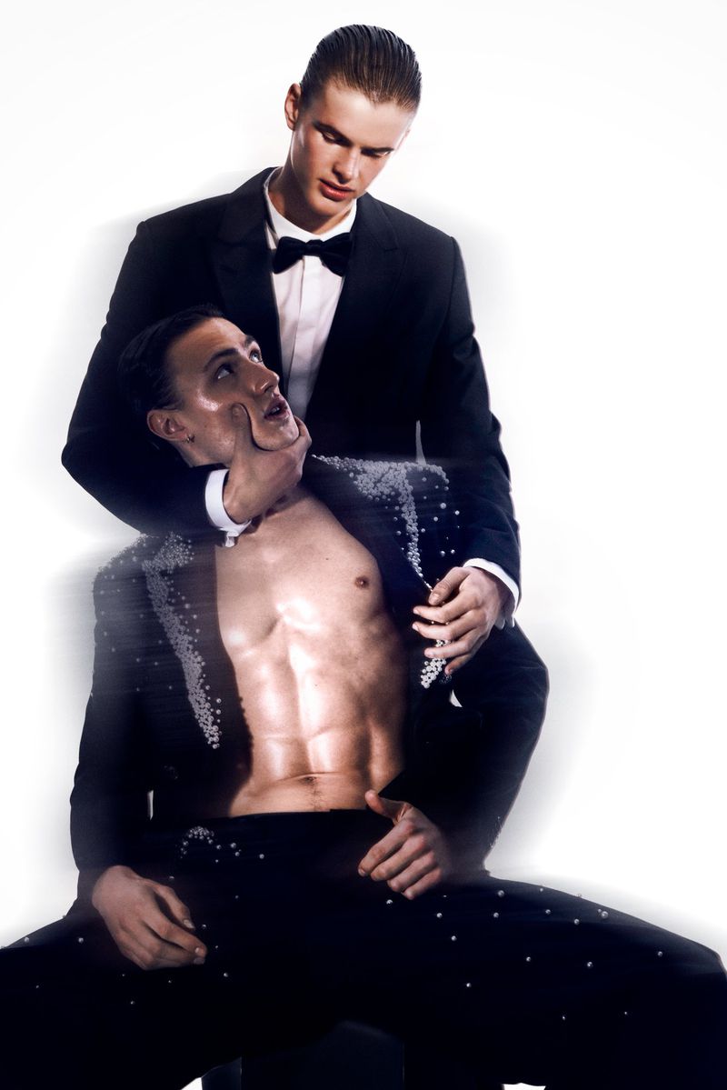 Two men in formal wear, one kneeling shirtless with an open jacket as the other stands behind him, hands near his neck, on white.